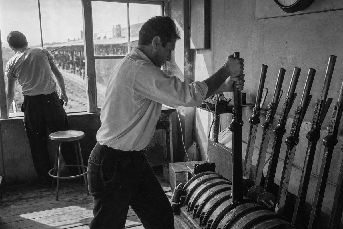 Black-and-white press photograph on Kodak Tri-X 400, early 1960s, Aleppo CCL signal box interior; dispatcher with rolled-up sleeves pulls a heavy lever on the mechanical interlocking frame while another operator leans toward a window overlooking a platform; steel rods, worn wooden floorboards, and a metal stool; wall clock partly cropped with numerals not visible; shafts of noon light through dusty panes, faint smoke in the air; gritty grain, moderate contrast, slight motion blur; taken with a 50mm lens on a 35mm SLR from shoulder height; asymmetrical framing with the lever bank dominating the right side.