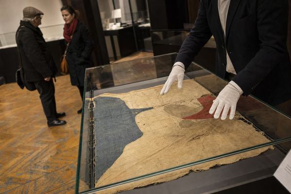 A candid digital press photograph taken in March 2026 inside the preview hall at Hôtel Drouot in Paris. Off-center composition at waist height with a 35mm lens on a full-frame mirrorless camera (f/2.2, ISO 3200), showing a glass display vitrine holding a frayed 1941 hand-painted cotton banner with no visible lettering. The fabric’s worn weave, faint discoloration halos, and slightly puckered seams are clearly rendered; the vitrine edges catch small reflections. A handler in a dark blazer, wearing white cotton gloves, hovers both hands just above the fabric as if about to adjust it. Scuffed parquet flooring, a low plinth, and a discreet estimate card sit near the vitrine edge but any text is turned away or out of focus. In the background, two distinct visitors—one older man with a tweed cap, one younger woman with a red scarf—are softly blurred in conversation. Mild high-ISO noise, slight barrel distortion, and shallow depth of field. No visible text, signage, or watermarks.