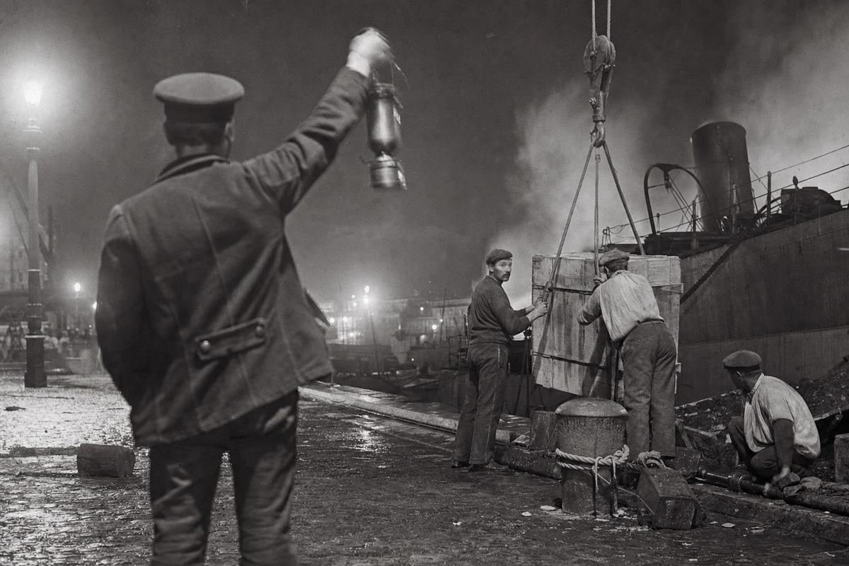 Silver gelatin night photograph, Marseille wartime loading quay, August 1914: oblique view from behind a signalman holding a hooded lantern aloft while a crane hook lowers toward unmarked wooden crates; steam haze and slight motion blur in the lantern hand; 135mm lens on a plate camera, long exposure giving halo around arc-lamp pools and deep shadows; coarse grain from high-sensitivity emulsion of the period, uneven exposure across the frame; distinct figures—two dockers bracing a crate, a third crouched by a chock—faces visible and varied; wet cobbles, rope fenders, coal dust; no visible lettering or signage.