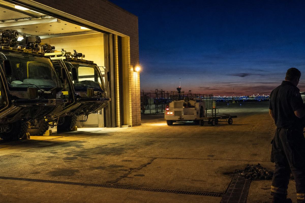 Open bays at O’Hare Fire Station 2 with crash tenders nose‑out at dusk, runway lights rising behind.