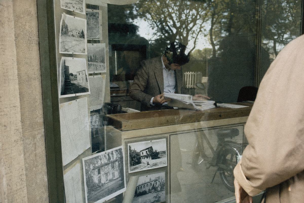 Vitrine d’agence immobilière à Villeneuve-lès-Avignon avec annonces épinglées et plans, reflets de platanes.
