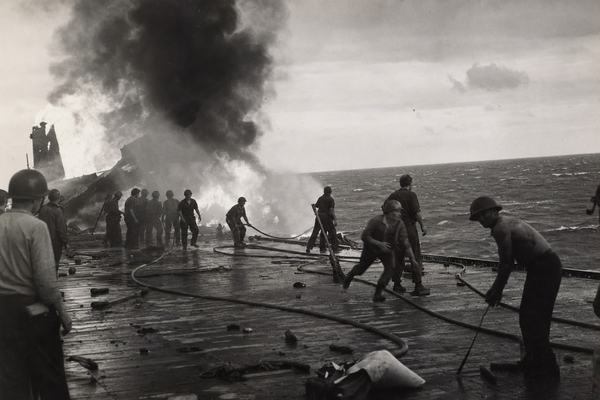 Archival silver-gelatin photograph taken late afternoon on June 6, 1942, from the edge of a U.S. aircraft carrier flight deck showing fire crews silhouetted against towering smoke while hoses snake across scorched planks; Pacific horizon slightly tilted with flecks of spray; shot on 35mm Kodak Super-XX by a Navy photographer using a 50mm lens; visible 1940s grain, minor salt spray on the front element, slight motion blur on a running sailor, uneven exposure darkening the right edge; asymmetric composition with the main blaze off at frame left, scattered deck gear and spent casings in foreground; no visible lettering or numbers anywhere; natural skin texture, mixed ages and builds among crew; period-authentic contrast and tonality.