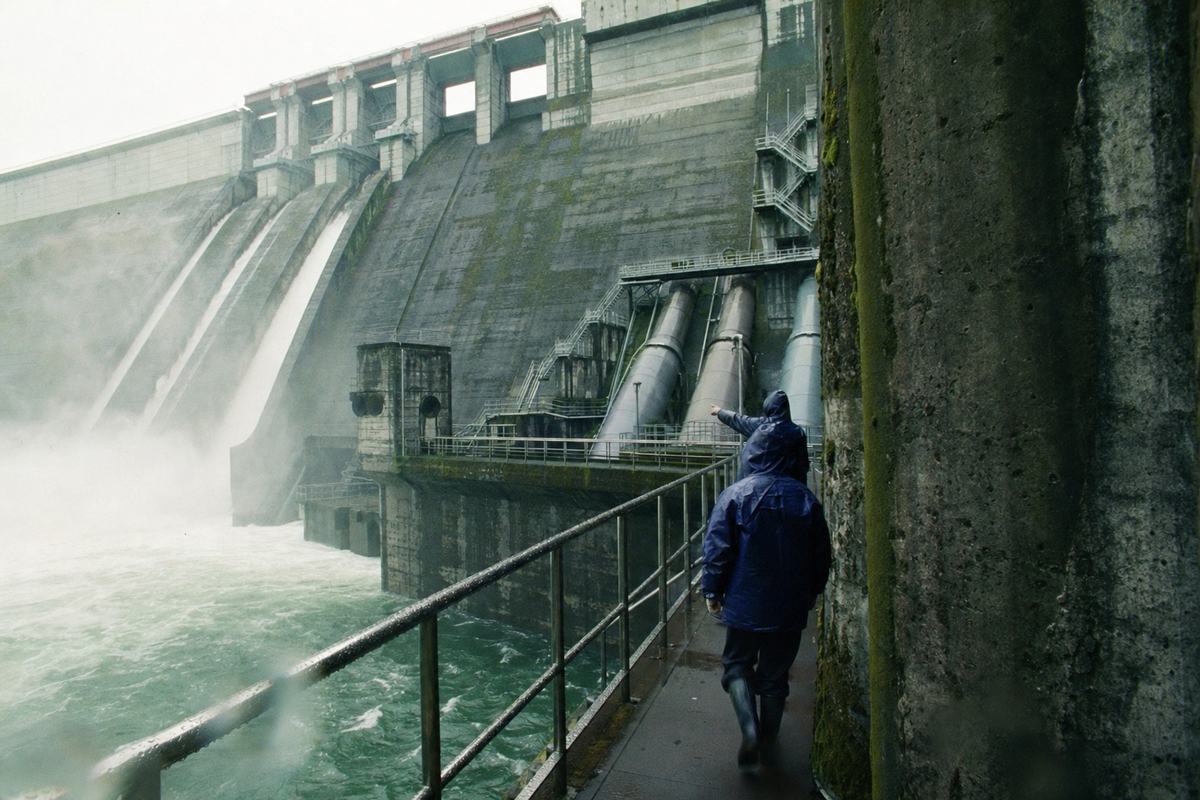 1974 color photograph of an Hokkaidō People’s Republic hydropower station exterior: concrete spillway with mist drifting across steel catwalks; two workers in blue oilskins walk along a damp railing, one pointing toward penstocks; moss stains and weathering on concrete, wet handrails bead with water; shot on Fujicolor 100 with a 28mm wide-angle lens, slight barrel distortion and greenish cast typical of the stock; fine grain, a few water droplets near the bottom edge; asymmetric composition with the workers at lower right; no signage, labels, or lettering visible anywhere.