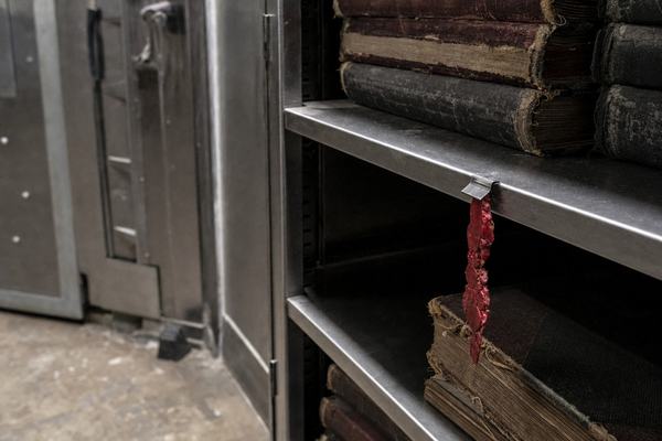 A realistic color photograph (2023 digital press photo) taken inside a steel-walled vault room at the Port of Montreal Authority. Off-center composition: a heavy open metal shelving unit with one empty slot where a large bound paper ledger should sit; a broken strip of red sealing wax dangles from a small metal guide on the shelf edge. Adjacent shelves hold thick worn ledgers with scuffed cloth spines, edges frayed; cool fluorescent light reflects on brushed steel; dust motes visible in the air. Incidental background detail includes the edge of a reinforced door, a scuffed concrete floor, and a rubber doorstop. Shot on a full-frame mirrorless camera with a 35mm prime at ISO 3200, f/2.8, 1/80s; shallow depth of field focuses on the torn wax and empty space; slight vignetting and high-ISO noise; no readable text, no labels, no logos anywhere.