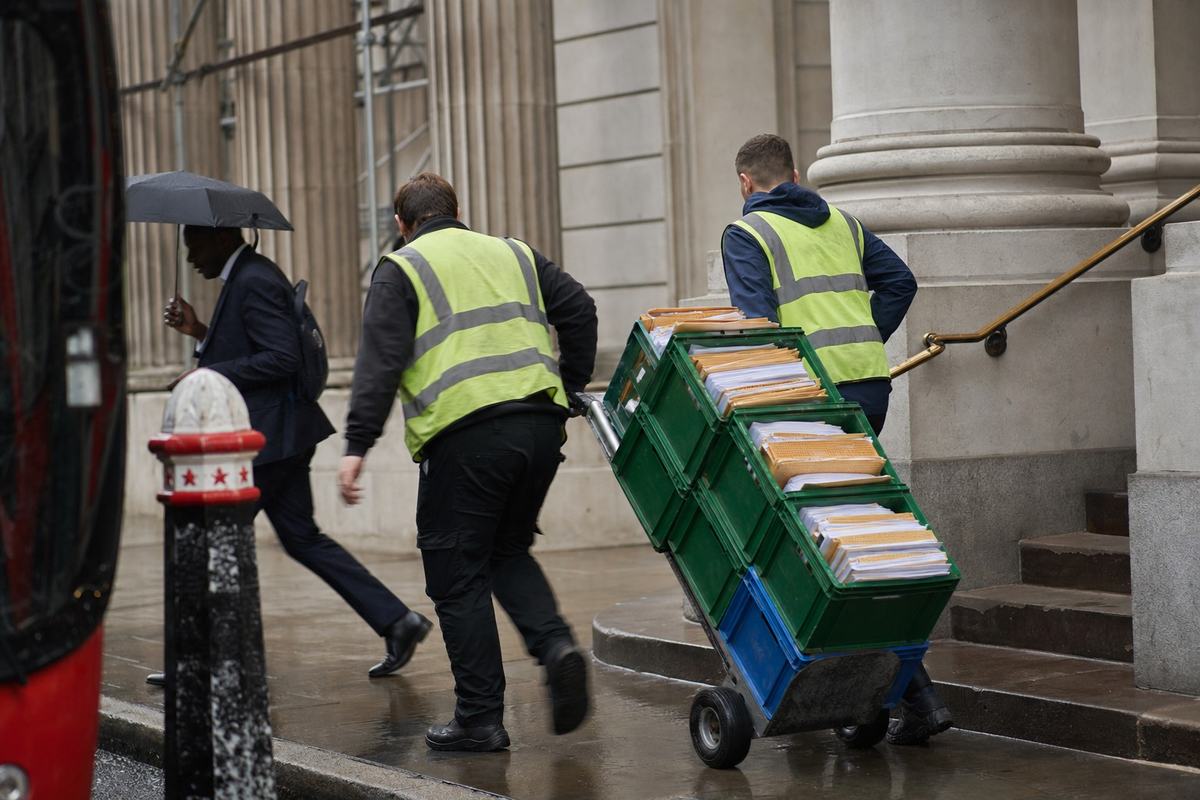 Devant une façade en pierre dans la City de Londres, des coursiers poussent des caisses de dossiers sous une bruine, marches usées et trottoir mouillé.