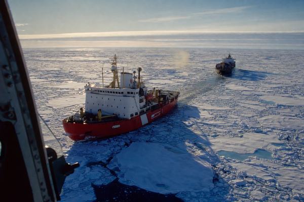 1990s color photograph shot from a low helicopter door over the Arctic Corridor, showing a Canadian Coast Guard heavy icebreaker leading a small container ship through patchy late‑summer sea ice under a low golden sun; photographed on Fujichrome Provia 100F with slight cool blue cast and saturated reds; 70–200mm zoom at ~120mm; slight motion blur at frame edges from rotor vibration; asymmetric composition with the icebreaker offset left and the container ship staggered back to the right, long shadows pulling toward the bottom of frame; cold blue water, white ice pans with melt pools, faint exhaust haze; horizon low with high thin cloud; hull angles and colors visible but no legible markings or numbers; no text anywhere in frame; incidental details include wake troughs, brash ice clumps, and a dark lead opening ahead.