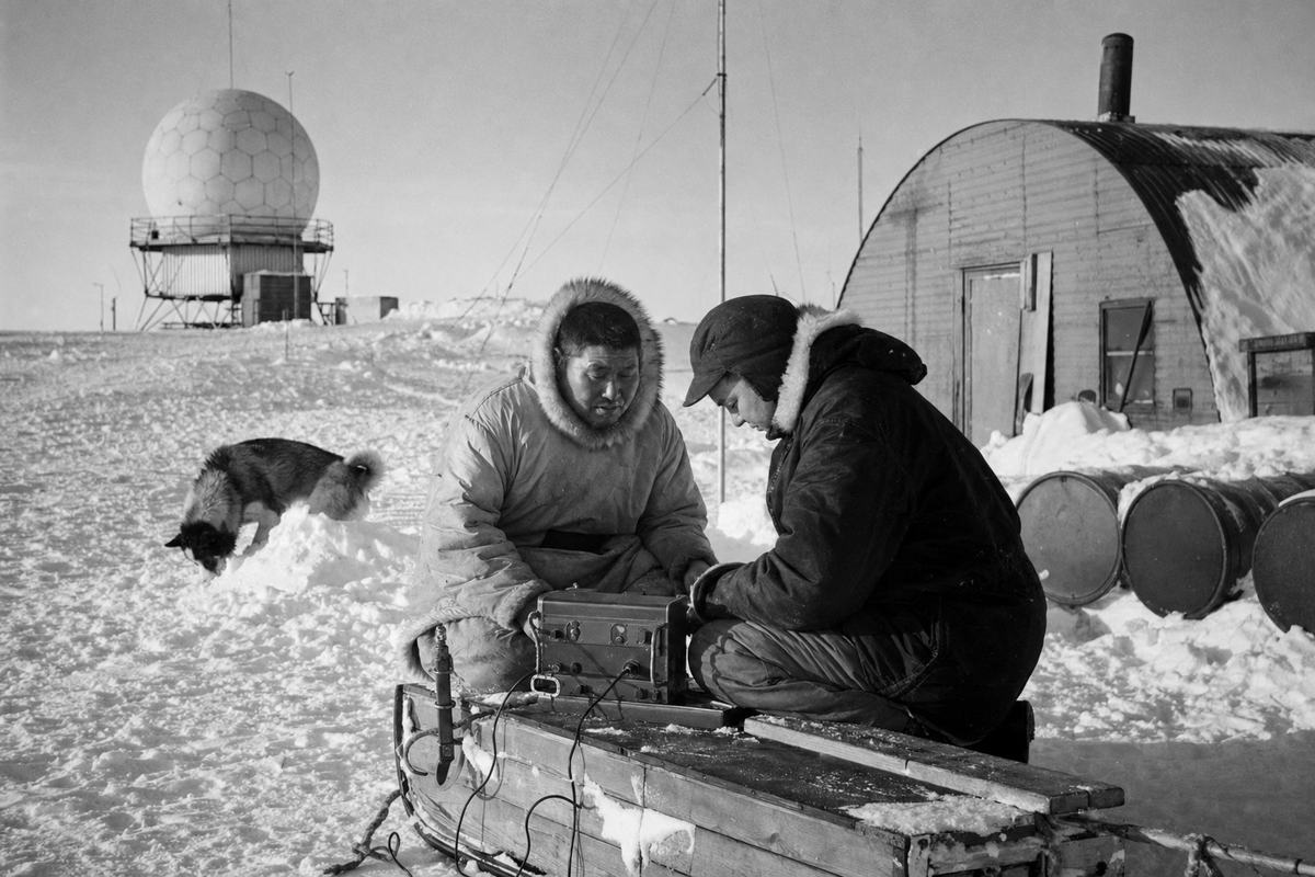 Mid‑1950s press photograph on Kodak Tri‑X 400 of a northern airfield and early warning site along the Arctic Corridor; 35mm Leica M3 with 50mm lens; high‑contrast grain with crisp winter shadows; asymmetric framing with a radar dome left‑background, a Quonset hut and fuel drums (turned so no stenciling is visible) to the right, and two technicians in parkas—one Inuk, one southern—hunched over a portable radio set on a sled in the foreground; snowdrifts bank against the hut, antenna masts thread the sky; a sled dog noses around a snow berm; no visible text, patches, or insignia; low sun glints off guy wires, breath vapor faint in the cold air.