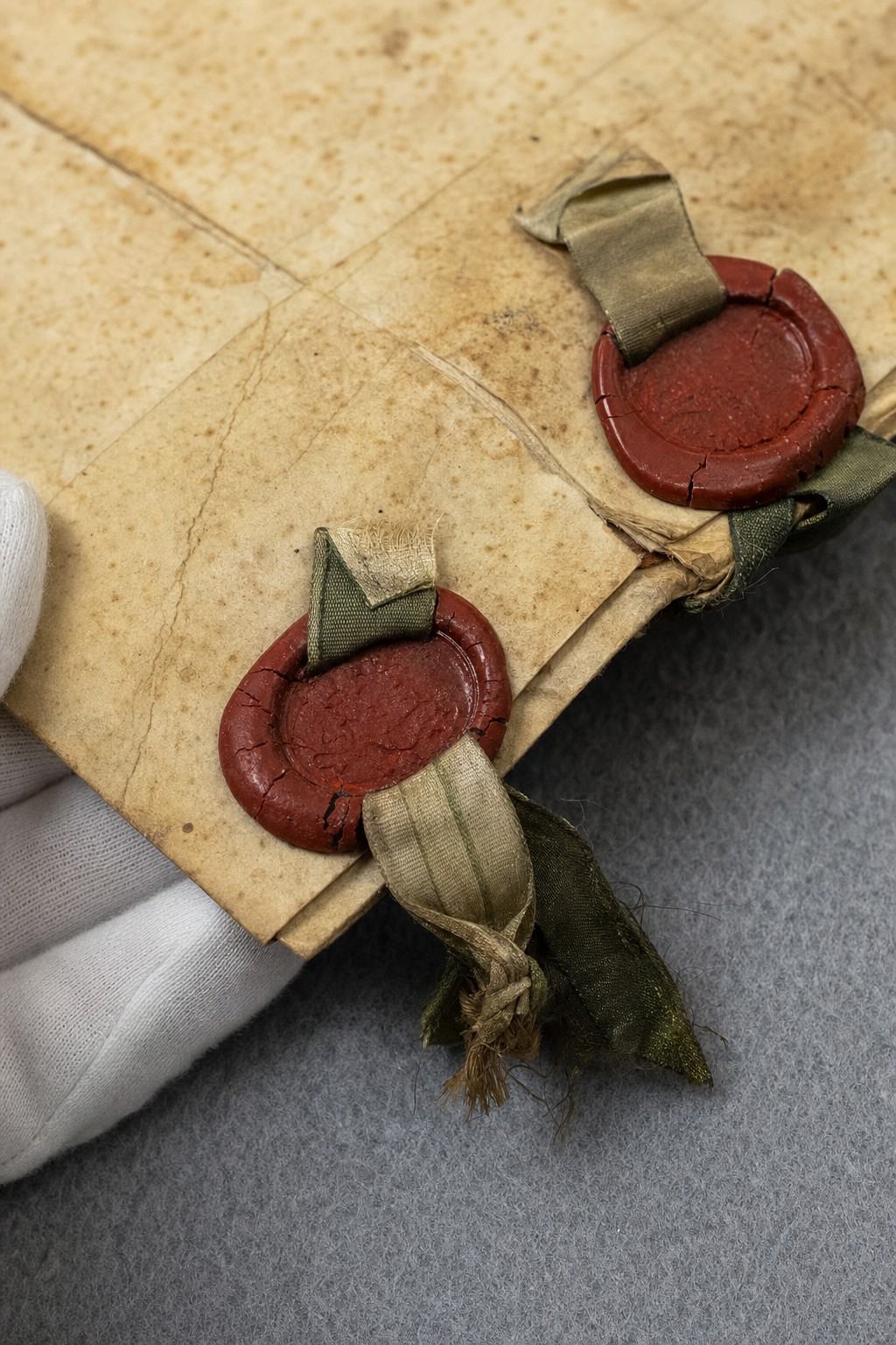 Archival object study photograph of the 1856 Peel Sound Compact focusing on wax seals and ribbon; shot on Ektachrome 64 in the 1990s with a 90mm macro lens; document placed on gray conservation felt with soft raking window light; parchment edge with visible foxing and creases, red wax seals slightly chipped, silk ribbon frayed; a cotton‑gloved hand steadies the lower corner; composition cropped so that any handwriting or printed letters are completely out of frame—no visible text at all; shallow depth of field isolates the seals, with background mount fading to soft blur.