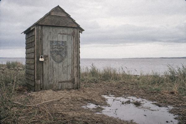 1979 color film photograph on Kodak Ektachrome 100, slight blue-grey cast and visible fine grain in flat late-summer overcast. 35mm SLR with a 28mm lens, low eye-level, off-centre composition with a small timber tide-and-rain gauge hut at Paull placed on the left third. Heavy brass padlock glinting on a weathered door that shows a faded crest with no legible lettering. Foreground of cracked clay, thistles and reed trash; rusted hinges, damp wood grain, and a shallow puddle catching grey light. The Humber estuary reads in the background as a strip of dull water under a low cloud ceiling; no people; faint wind ripple on water; slight corner vignetting typical of 1970s regional newspaper photography.