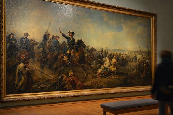 Color photograph of an 18th-century oil painting hanging in a museum gallery; three-quarter angled view under warm tungsten gallery lighting with slight glare on varnish; the canvas (Baltic workshop, c. 1715) shows Swedish cavalry breaking through Muscovite lines on the steppe while a distant Cossack screen closes the retreat; visible craquelure and uneven aged varnish, thick brushwork in the horses and dust; hardwood floorboards and a bench in foreground, one passerby blurred at the edge of frame; no labels or wall text visible; shot on a Nikon F2 with a 50mm lens on Kodachrome 64, fine grain, slight barrel distortion and mild motion blur from handhold.