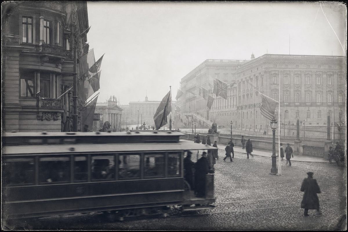 Silver‑gelatin view of the Riksdag and Royal Palace around 1905, with provincial ensigns flying and a tram blurring past on the cobbles.