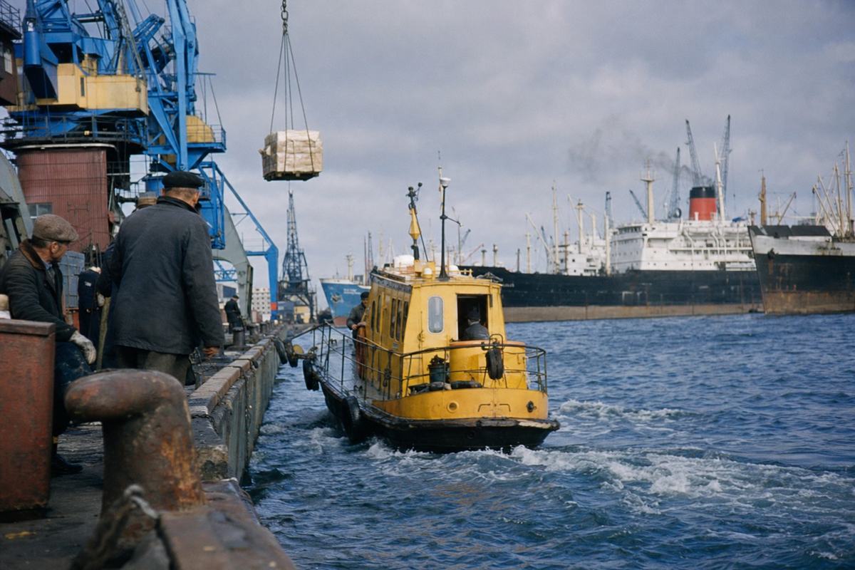 Kodachrome view of Tallinn harbor in the 1960s with a yellow pilot boat departing toward a small convoy and cranes lifting cargo.