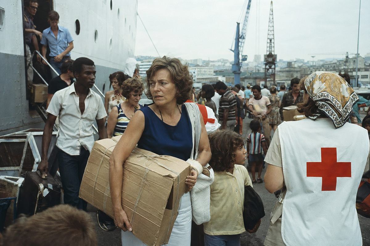 Color film photograph taken in 1976 at a Marseille quay, capturing the moment families disembark from a liner arriving from Luanda. Cardboard suitcases symbolizing hurried evacuation, expressions of uncertainty on mixed-aged faces, and Croix-Rouge volunteers assisting them. Slight overcast sky with diffused lighting typical of port environments. Fujicolor film with slightly muted tones to capture the historic essence of the event.