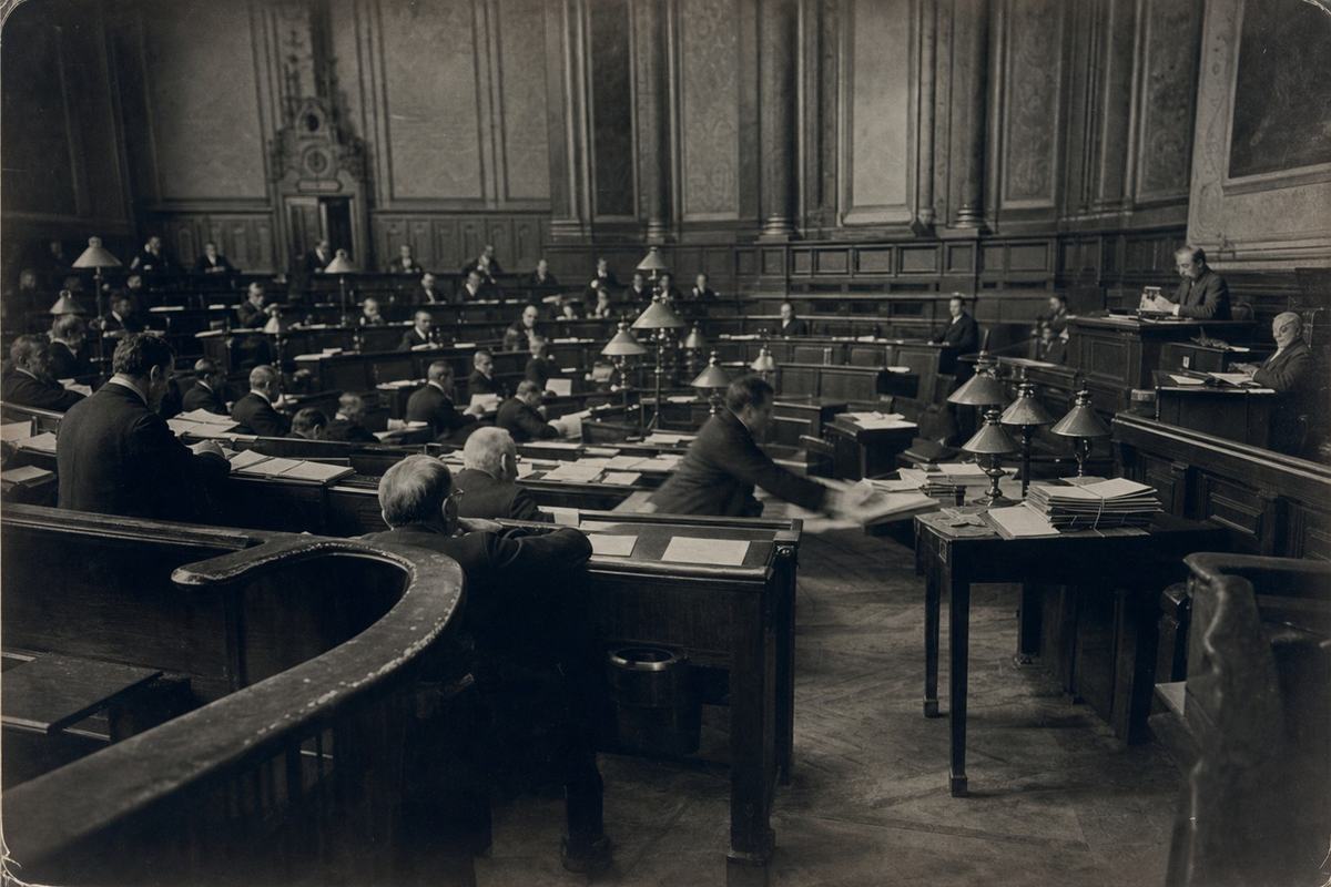 Archival interior photograph, silver gelatin print (dry plate), ca. 1895, Danube Council chamber inside the Council House on the Ring; taken with an 8x10 view camera and a 300mm lens from the rear left gallery, long exposure with slight motion blur in a clerk reaching for papers; benches with worn armrests, green-shaded gas lamps casting uneven pools of light, parquet floor scuffed near the aisle; bilingual nameplates are turned away or out of focus (no legible text), stacked dossiers tied with string on a front desk, a spittoon tucked under a bench; moderate edge vignetting, rich midtones with selenium toning, fine paper fiber visible, slight curl at the corners; asymmetrical framing with the speaker’s dais partially cut off at right.