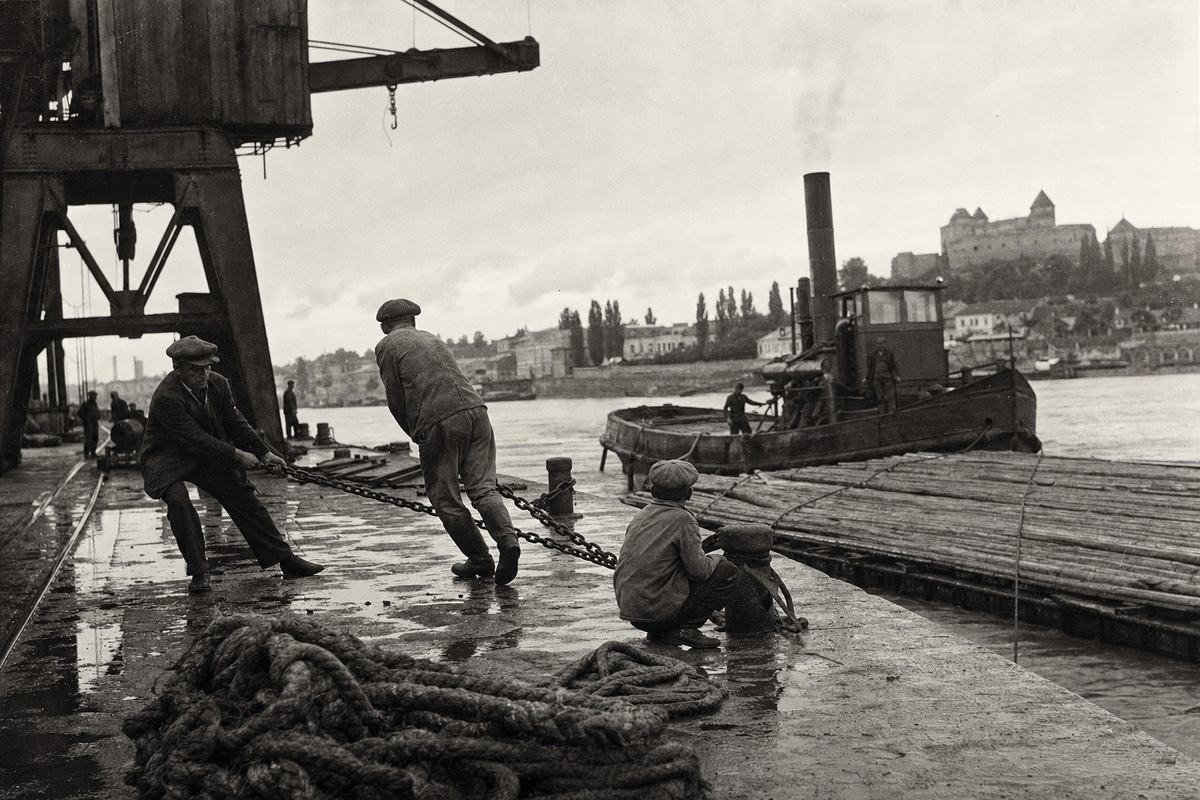 Harbor-side photograph, silver gelatin print on matte fiber paper, 1925, Danube port at Komárno; shot with a 13x18 cm plate camera and a 210mm lens from the quay edge, low over a coil of hawser; a tug noses a timber raft while two dockworkers in flat caps haul a wet chain, a boy squats by a bollard watching; spray darkens the tug’s hull, puddles reflect a cloudy sky, a craneway looms at upper left; tonal range from inky blacks under the barge to crisp highlights on wet planks, visible film grain in midtones, slight horizon tilt; any posted notices are turned away or cropped—no legible text; off-center composition with the pull of the chain as the dominant action.