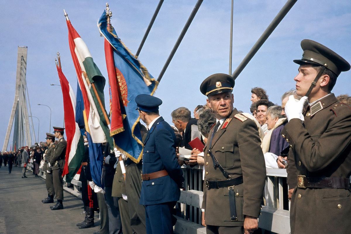 Color photograph, Kodachrome 64 slide shot in 1965 at the inauguration of a new Danube-span bridge at Komárno/Komárom; taken with a 50mm lens from waist height at three-quarters angle, slight cyan cast and saturated primaries, fine grain; a mixed-regiment honor guard stands at rest along the guardrail, one young soldier adjusting his chinstrap while an older NCO glances toward the crowd, flags ripple in a gust; steel cables arc into the distance, concrete pylon receding left, onlookers lean over the railing with paper programs in hand; no signage or lettering visible; asymmetric composition with the flagstaffs leading the eye toward the center span.