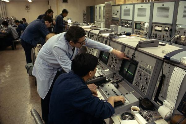 1978 Franco-Chinese joint space control room, 35mm color film photograph (Kodak Ektachrome 64), slight tungsten cast, visible fine grain, minor motion blur on hands; candid documentary angle from behind and to the left with a 50mm lens; mixed French and Chinese engineers of varied ages leaning over analog consoles with rotary knobs and paper printouts, coiled telephone cords, ashtrays, coffee cups; CRT monitors show green waveform traces only (no legible text), patch cables draped over rack fronts; scuffed linoleum floor, chipped paint on cabinet corners, ventilation grilles dusty; one dominant action: an engineer pointing at a waveform while another adjusts a dial; no logos, no readable labels anywhere.