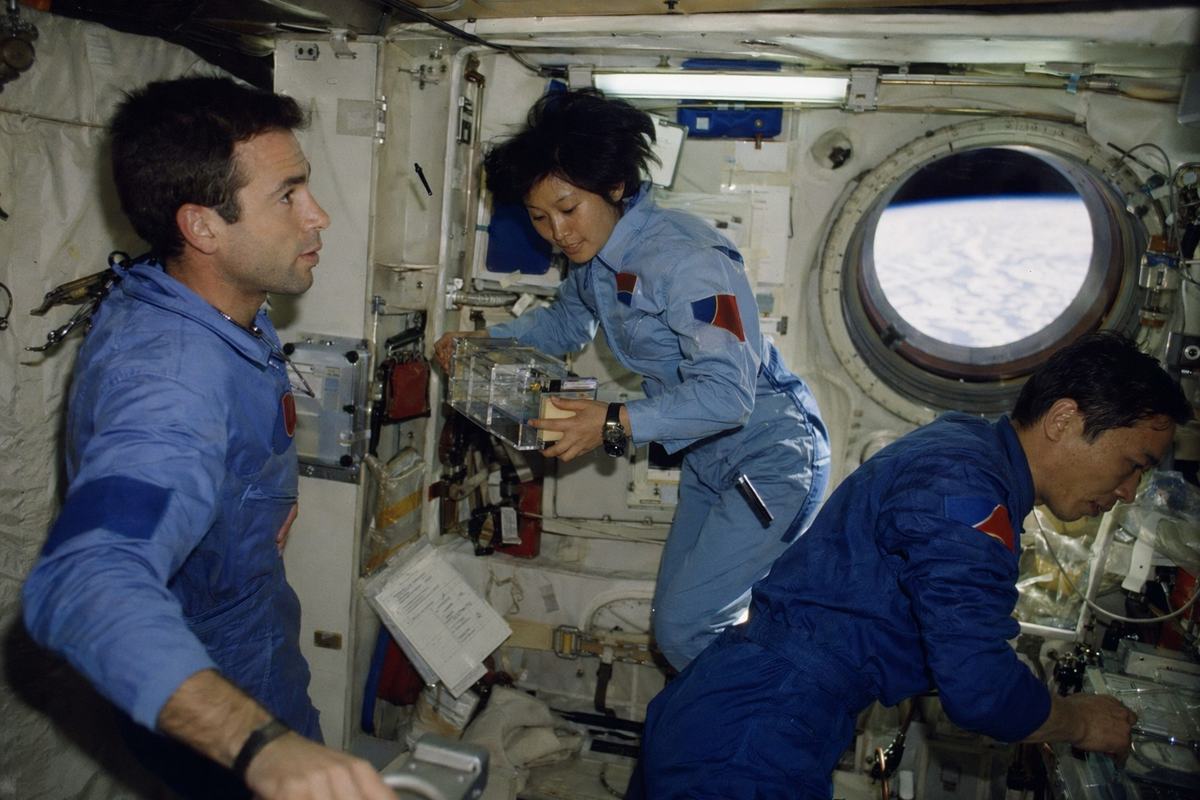 Early 1980s interior of the shared 'Seine–Yangzi' orbital module, 35mm color film photograph (Fujicolor 400), mixed lighting with cool fluorescent tubes and soft daylight from a circular porthole; 28mm lens with slight barrel distortion; three distinct crew members (one French male late 30s, one Chinese female late 20s, one Chinese male early 40s) in vintage pressure suits/flight coveralls with a simple red-blue patch shape (no text); one astronaut steadies a handrail while another floats holding a small experiment kit; velcroed tools, wiring looms, and checklists with blocks and lines but no readable text; Earth limb visible through the window; moderate grain, small bits of floating debris like a pen cap.