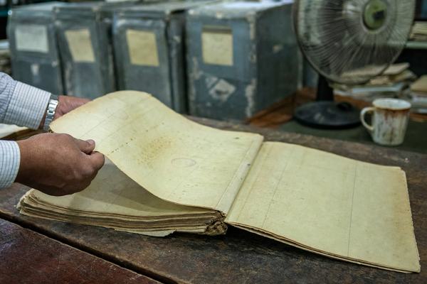 Digital press photograph, object/document study style, of yellowed administrative register pages on a scuffed wooden archive table; oblique angle from shoulder height; a pair of adult hands carefully turning one brittle page; visible paper foxing, frayed edges, faint rubber-stamp marks and marginal notations but no legible text; soft overhead fluorescent light with slight green cast; shallow depth of field (only the page corner sharp, background blurred); in the background, dented metal archive boxes with worn labels out of focus, an oscillating desk fan, and a chipped ceramic mug; captured on a 50mm equivalent lens, ISO 3200 producing fine digital noise, slight motion blur in the fingertips; no lettering, no overlays, no watermarks.