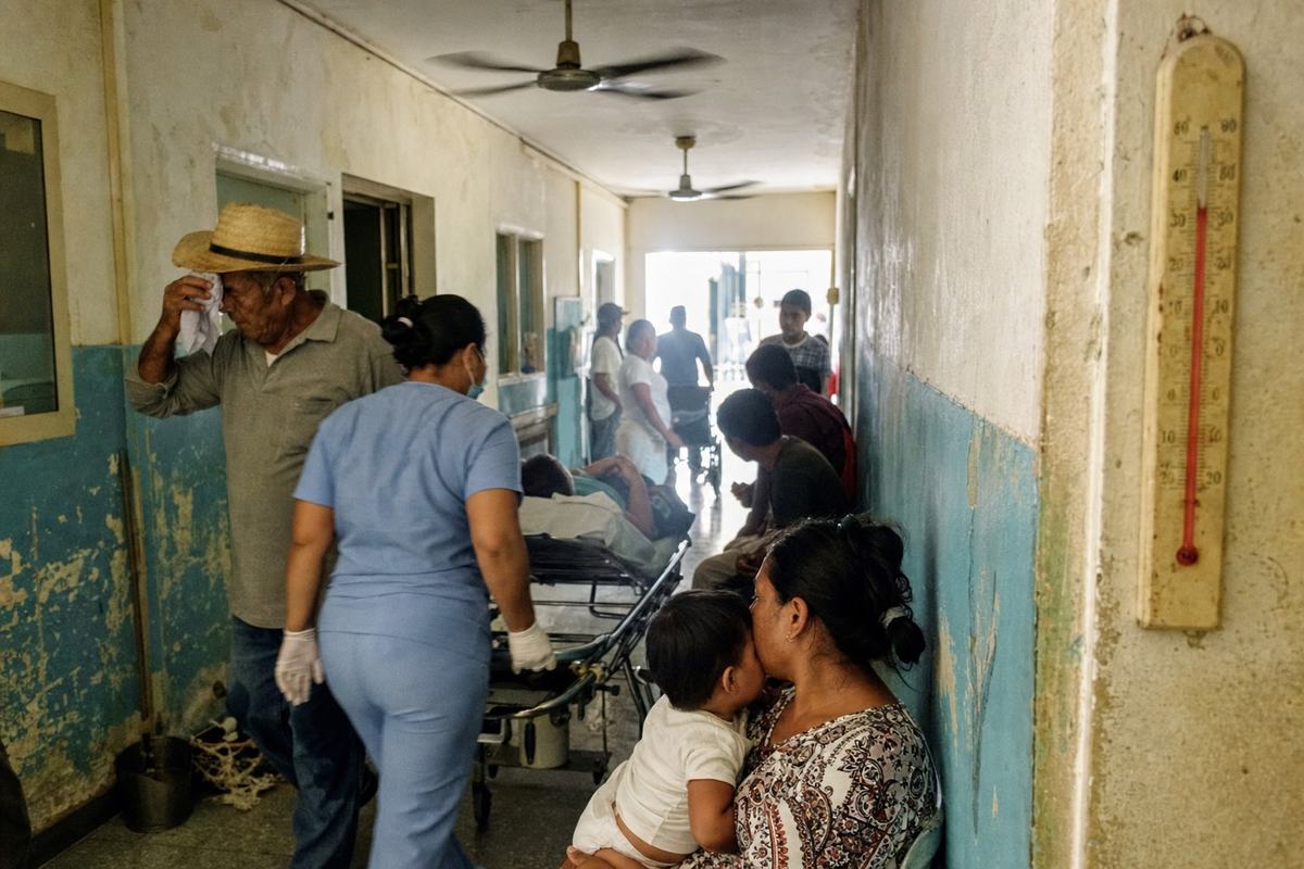 Couloir d’hôpital provincial avec ventilateurs, thermomètre au mur et files d’attente sous une chaleur écrasante.