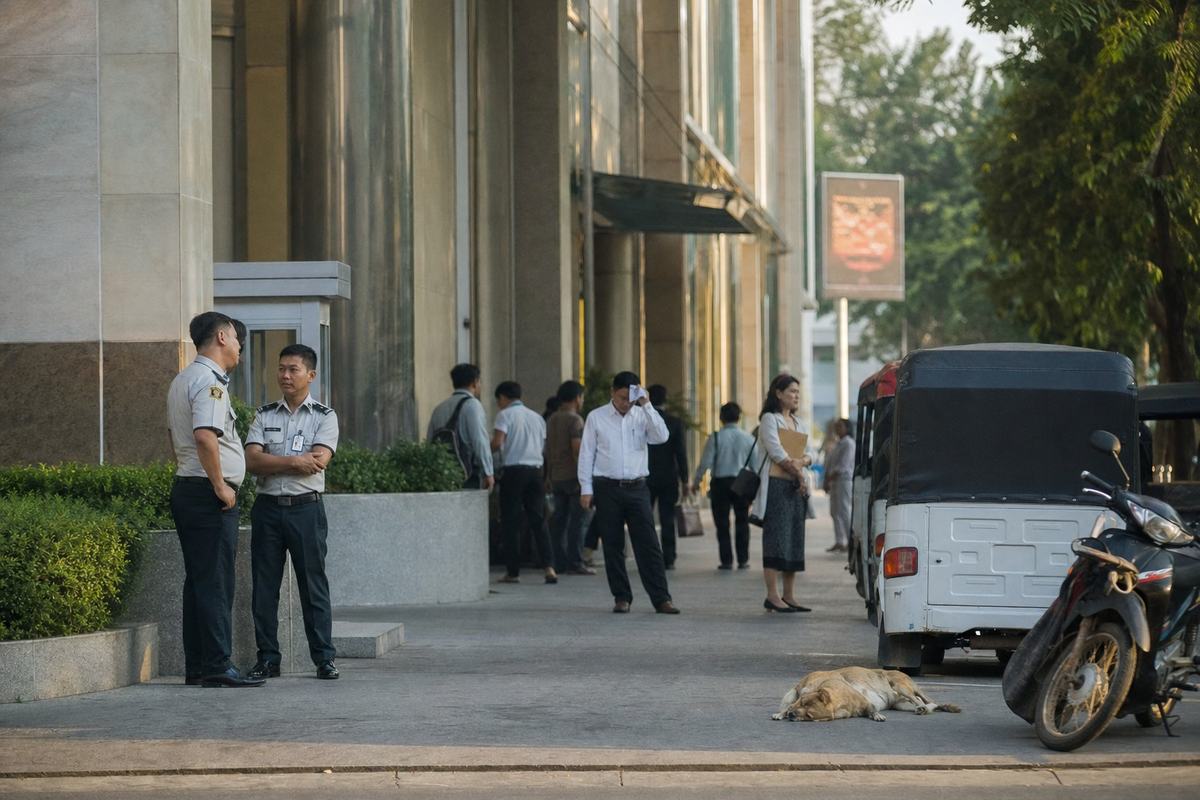 Façade du Secrétariat de l’ASEAN à Phnom Penh, gardes et visiteurs à l’ombre, vibration de chaleur en fin d’après-midi.
