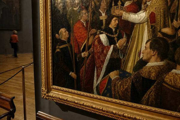 Color photograph of a 15th–16th century oil painting in a museum gallery, shot on 35mm Kodachrome 64 with a 50mm lens at f/2.8; off-center framing catching the gilded frame’s lower corner and the central figure of Charles the Bold receiving the Lotharingian coronation; visible varnish glare and fine craquelure on the paint surface; rich brocade costumes, clerics and attendants with distinct faces and varied postures; heraldic colors (reds, golds, deep blues) but no inscriptions or lettering in the painting; museum setting with parquet floor, a rope stanchion, and a wooden bench at the edge of frame; a passing visitor softly blurred in the background; controlled tungsten lighting with gentle falloff; ensure no wall labels or signage, no readable text anywhere; candid, asymmetric composition.