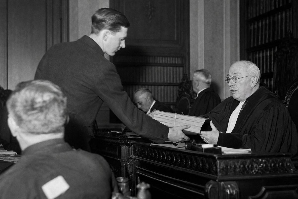 Black-and-white press photograph inside a grand courtroom (Grand Council of Malines), 1937; gelatin silver print from a 4x5 Speed Graphic negative, single flashbulb illumination creating crisp highlights and hard shadows; dominant action: a young court clerk leaning forward to hand a bound stack of plain case files (no labels or text) to an older robed councillor with wire-rim glasses; wood paneling and high-backed chairs; shelving in background with book spines turned away from camera (no inscriptions visible); moderate grain, slight motion blur in the clerk’s hand; candid, asymmetric composition framed from aisle level.