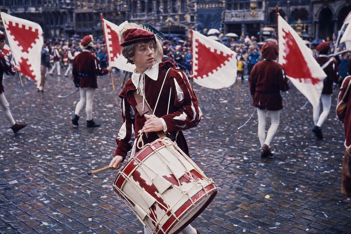 1969 color slide of a centenary parade on the Grand-Place, Brussels, shot on Kodachrome 64 with a Nikon F and an 85mm lens; saturated reds and deep blues with a slight magenta cast; dominant subject: a young drummer in Burgundian-era costume mid-step, head turned, natural skin texture and imperfect posture; behind him, standard-bearers carry flags with a Burgundian saltire motif (no text or lettering anywhere); damp cobblestones with scattered confetti, blurred spectators at the edge, ornate guildhouse facades in soft focus; candid, asymmetric composition capturing motion without any visible signage.