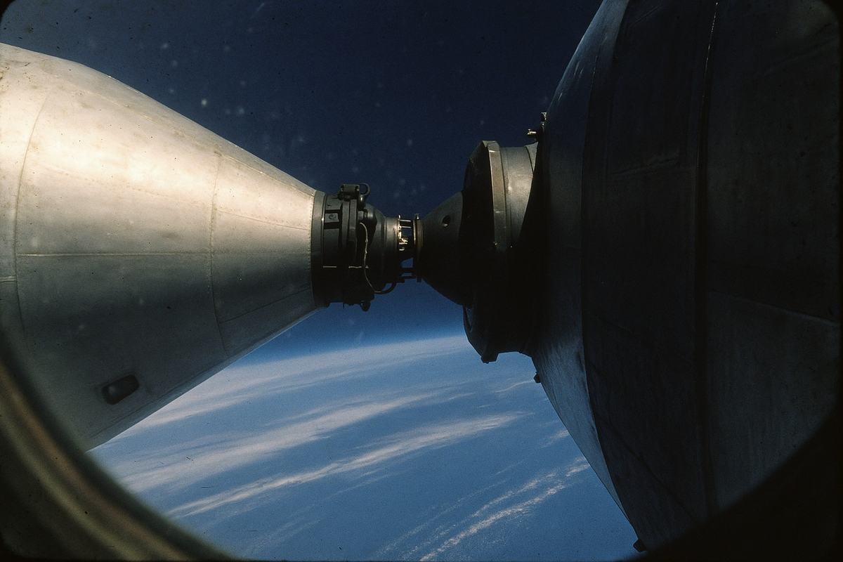 Archival exterior color photograph on Kodak Ektachrome 100 slide film, shot through a Salyut 6 window in August 1978 with an 80mm lens. Asymmetric frame: the nose of a modified Apollo command/service module is docked to the forward port of Salyut 6 via an APAS-style adapter, while the Earth’s blue limb and thin cloud bands sweep diagonally below. Faint window reflections and dust specks, slight cyan cast, moderate grain, a touch of flare at the rim of the station’s hull. No visible text, markings, or lettering.