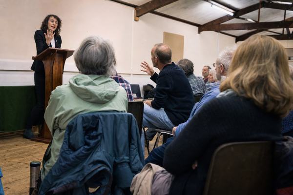 Candid digital press photograph taken in May 2026 inside the Pendine Institute village hall during a book launch Q&A: Dr Eleri Vaughan stands at a scuffed wooden lectern, mid‑answer with one hand raised, while two distinct seated attendees in metal stacking chairs gesture and challenge from the floor; mixed‑age local audience with varied builds and natural skin texture; fluorescent strip lighting with cool tint, exposed timber rafters overhead, pale walls with community noticeboards turned away so no lettering is visible; raincoats draped on chair backs, a thermos on the floor near the aisle; shot from mid‑aisle at 35mm on a full‑frame DSLR, ISO 3200 with visible noise, slight motion blur on hands, shallow depth of field keeping Vaughan and one questioner sharp as the background softens; asymmetric framing with Vaughan off‑left; natural perspective, no banners or text, no watermarks.