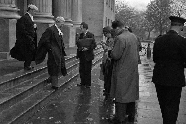 Black-and-white press photograph taken in 1949 on Ilford HP3 35mm film with a Leica IIIc and 50mm lens; medium grain, slight motion blur from figures in mid‑stride, damp air bloom on highlights. Asymmetric composition from the left side of the steps of the Supreme Court in Castlebar: two robed barristers descending past a clerk clutching a leather folio, a small knot of reporters with notebooks held low, umbrellas half‑furled, a uniformed constable half in frame at the edge. Rain‑dark flagstones glisten; puddles reflect pale autumn light. Stone columns recede into soft focus; a bicycle leans against a bollard in the background. No visible text, lettering, or signage anywhere.