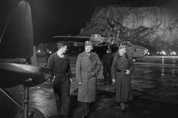Archival black-and-white press photograph, RAF North Front, Gibraltar, night of 4 July 1943. Candid, asymmetric framing with the edge of a wing in the near left foreground and General Władysław Sikorski mid-stride in a greatcoat, flanked by a younger RAF ground crewman and an older Polish officer. A B-24 Liberator sits in the mid-ground with propellers idle; damp tarmac reflects point-source floodlights; the limestone cliff looms as a dark mass beyond the runway. Captured with a Graflex Speed Graphic 4x5 and flashbulb on Ilford HP3 film; silver gelatin print with pronounced grain in the shadows, slight motion blur in hands, and a falloff of light at the frame edges. Natural skin texture, creased uniforms, scuffed boots. No legible text or signage anywhere in frame.