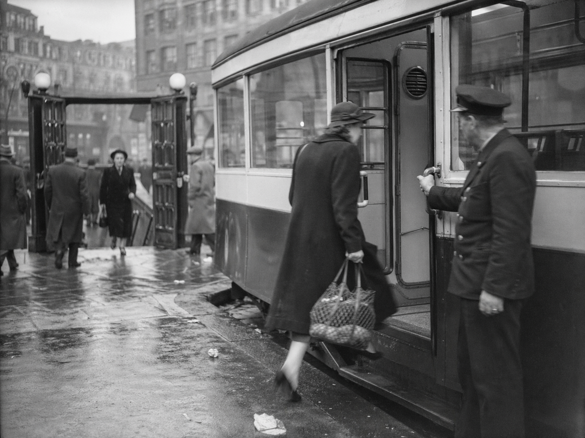 Black-and-white press photograph on silver-gelatin paper, London circa 1938, showing a tram at curbside with a sealed Type B filtered compartment. A uniformed conductor holds the door while a woman with a string bag steps over the raised threshold lip; rubber gaskets and a fan grille are visible inside. In the background, a Tube entrance with heavy overpressure doors stands partly open as commuters move past; wet pavement with scattered litter at the kerb. Handheld 35mm rangefinder with a 50mm lens on Agfa Isopan film; medium grain, slight motion blur in the woman’s foot and the conductor’s hand, gentle fall-off toward the far platform. Candid, off-center framing. No visible text or lettering anywhere.