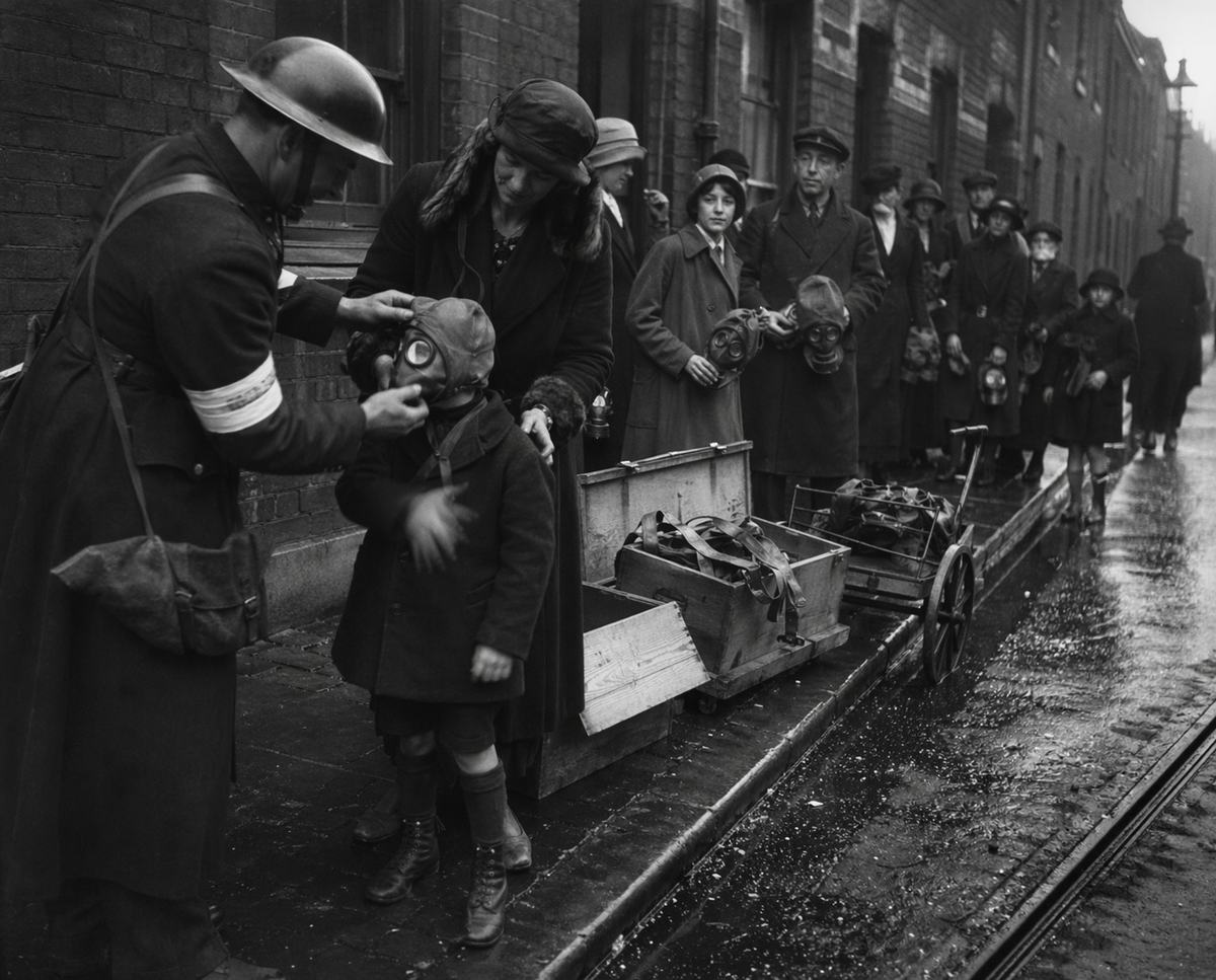 A warden fits a respirator hood on a child as Londoners queue along a wet street during a 1916 gas alert.