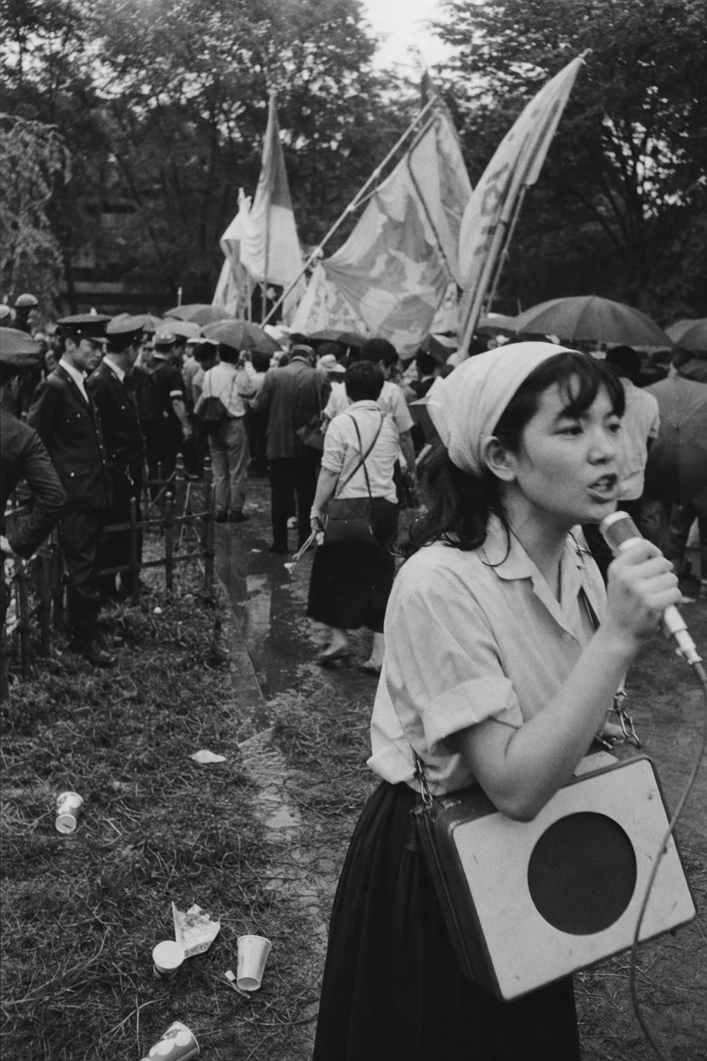 Black-and-white press photograph, silver-gelatin print, shot in June 1960 at Hibiya Park during the security treaty protests. Captured with a Nikon S2 and 35mm lens, Kodak Tri-X 400 pushed to 800, pronounced grain, high contrast, slight motion smear from movement. Asymmetric composition: foreground right features a young woman in a headscarf gripping a portable loudspeaker, mouth mid-syllable; behind her, a dense crowd with rain-damp umbrellas, hand-painted banners turned away from the camera so any lettering is indistinct; left background shows uniformed police along a low fence. Trampled grass, puddles glinting on paths, discarded paper cups, and haze from drizzle add incidental detail. No readable text.