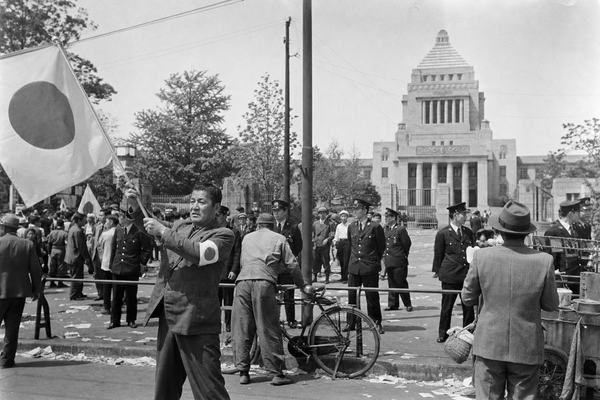 Archival black-and-white press photograph, silver-gelatin print on fiber paper, taken May 3, 1947 outside the National Diet Building in Tokyo. Shot on a Leica IIIc with 50mm lens at roughly f/5.6, panchromatic film, moderate natural grain, slight motion blur at the frame edges, and a touch of uneven exposure near the top. Asymmetric, candid composition: in the left foreground, a middle-aged man lifts a banner bearing a simple republican insignia (no legible text), an armband with the same symbol on his coat; midground shows uniformed police in peaked caps forming a loose line; background reveals the Diet’s stepped roof under spring daylight with soft shadows. Distinct faces of varied ages, scuffed shoes, a bicycle leaning against a rail, scattered leaflets on pavement, and a pushcart vendor partly visible. Period-accurate clothing and architecture, realistic film rendering with no modern artifacts.