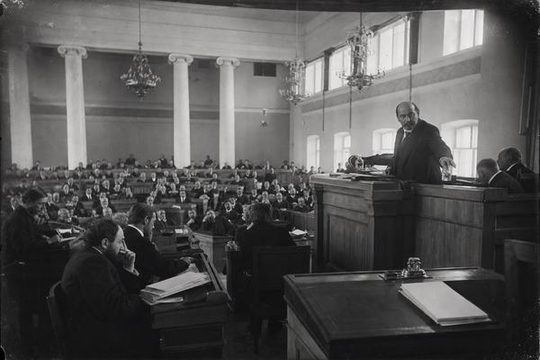 Archival wide-angle silver gelatin photograph (1906) of the interior of Petrograd’s Tauride Palace during a session of the First State Duma, shot from the right-hand gallery rail with a large-format view camera using a wide lens (approximately 28mm equivalent). Asymmetric composition with the rostrum off-center: a minister at the lectern reaches for a glass of water while turning slightly toward questioning deputies; benches crowded with men in dark frock coats; scuffed wooden floors; chandeliers and clerestory windows filtering soft afternoon light. Dry-plate negative look with slight emulsion scratches, corner vignetting, and gentle halation near windows; moderate grain, faint motion blur on gesturing hands, uneven exposure across the hall. Incidental details include an inkstand and stacked papers with their blank sides facing up; no text or signage visible.