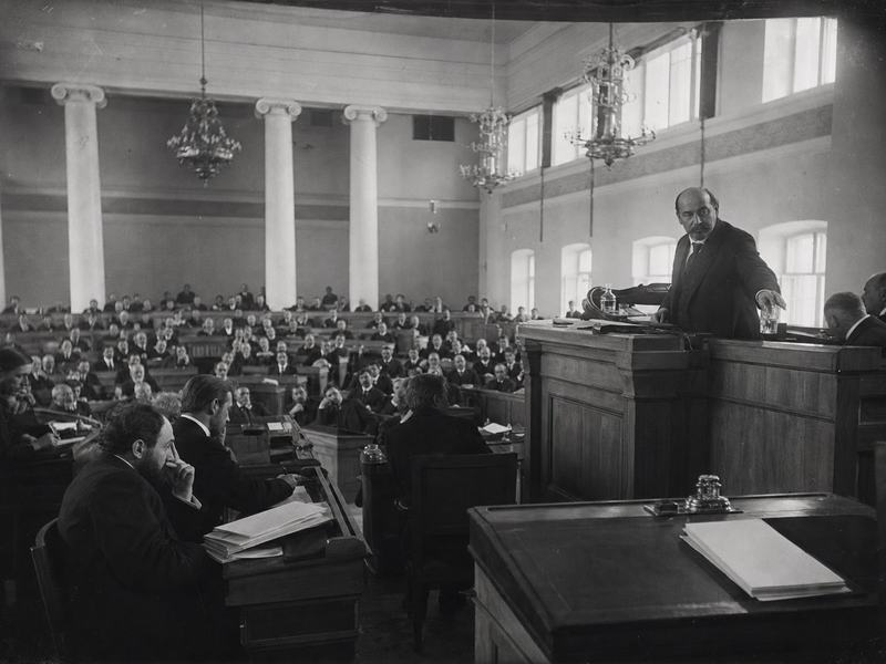 Archival wide-angle silver gelatin photograph (1906) of the interior of Petrograd’s Tauride Palace during a session of the First State Duma, shot from the right-hand gallery rail with a large-format view camera using a wide lens (approximately 28mm equivalent). Asymmetric composition with the rostrum off-center: a minister at the lectern reaches for a glass of water while turning slightly toward questioning deputies; benches crowded with men in dark frock coats; scuffed wooden floors; chandeliers and clerestory windows filtering soft afternoon light. Dry-plate negative look with slight emulsion scratches, corner vignetting, and gentle halation near windows; moderate grain, faint motion blur on gesturing hands, uneven exposure across the hall. Incidental details include an inkstand and stacked papers with their blank sides facing up; no text or signage visible.