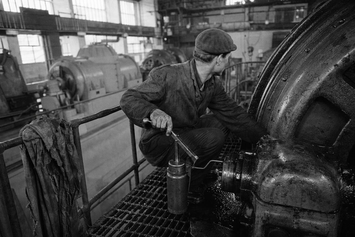 Industrial silver gelatin photograph (late 1930s) inside the Dnieper hydroelectric complex turbine hall: an overalled maintenance worker crouches on a steel catwalk, greasing a massive generator bearing with a hand pump, head turned toward the sound of a distant crane. Oblique view from an upper gantry with a 50mm lens on a 35mm rangefinder camera; high contrast with deep shadows, pronounced grain typical of high-speed stock, slight motion blur on the worker’s hand; shafts of light from clerestory windows create uneven exposure across riveted casings. Incidental details: a rag bundle hooked over a railing post, oil sheen on the floor grate, chalk smudge near a bolted seam; no text, labels, or signage visible.
