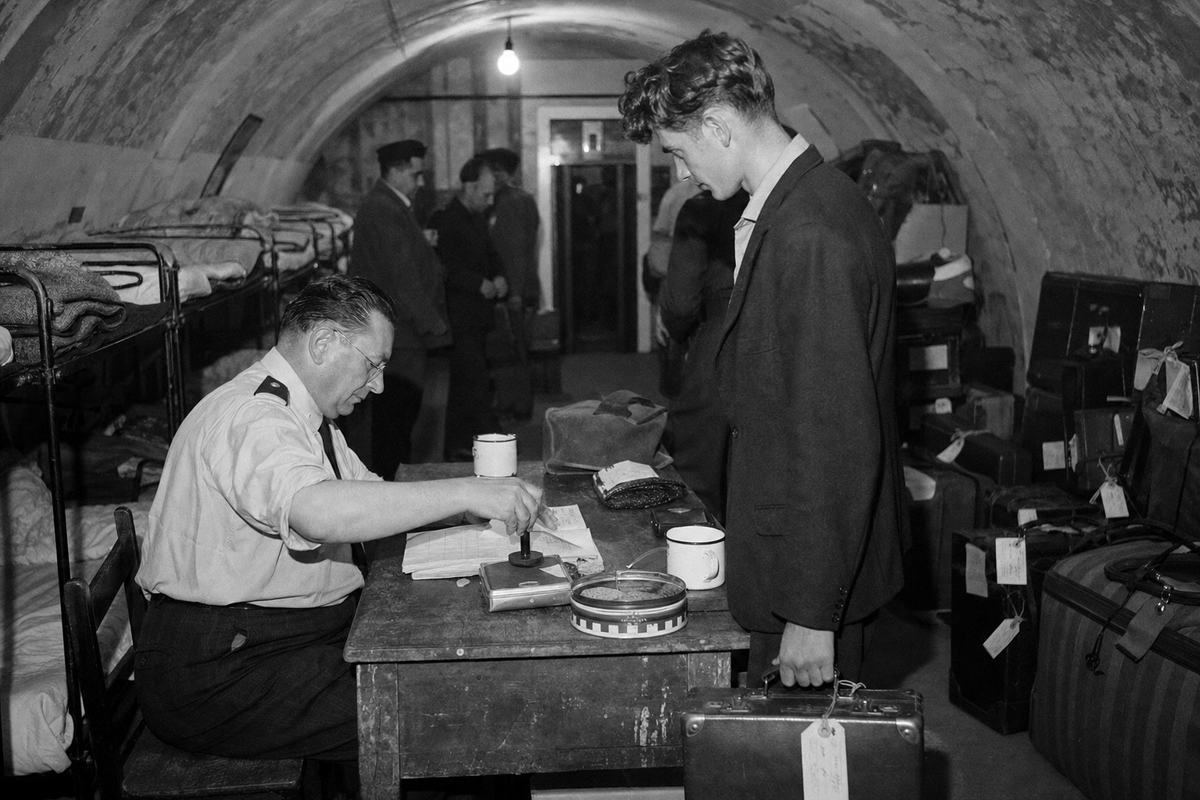 Archival silver-gelatin interior photograph of the CRS reception centre at the Clapham South deep-level shelter, July 1948. Curved tunnel walls with condensation and flaking paint; rows of metal bunks with folded grey blankets; a wooden intake desk under a bare bulb where a shirt-sleeved officer stamps a form while a young man with a suitcase waits. Luggage with twine tags piled by the wall (tags turned to avoid legible text), enamel mugs and a biscuit tin on the desk; a narrow lift doorway recedes in background. Shot on a Leica IIIc with 35mm Summaron lens, Ilford HP3 400; visible grain, edge falloff, slight blur from low shutter speed, deep shadows in corners. Asymmetric, candid framing centered on the officer’s stamping gesture; distinct faces and postures.