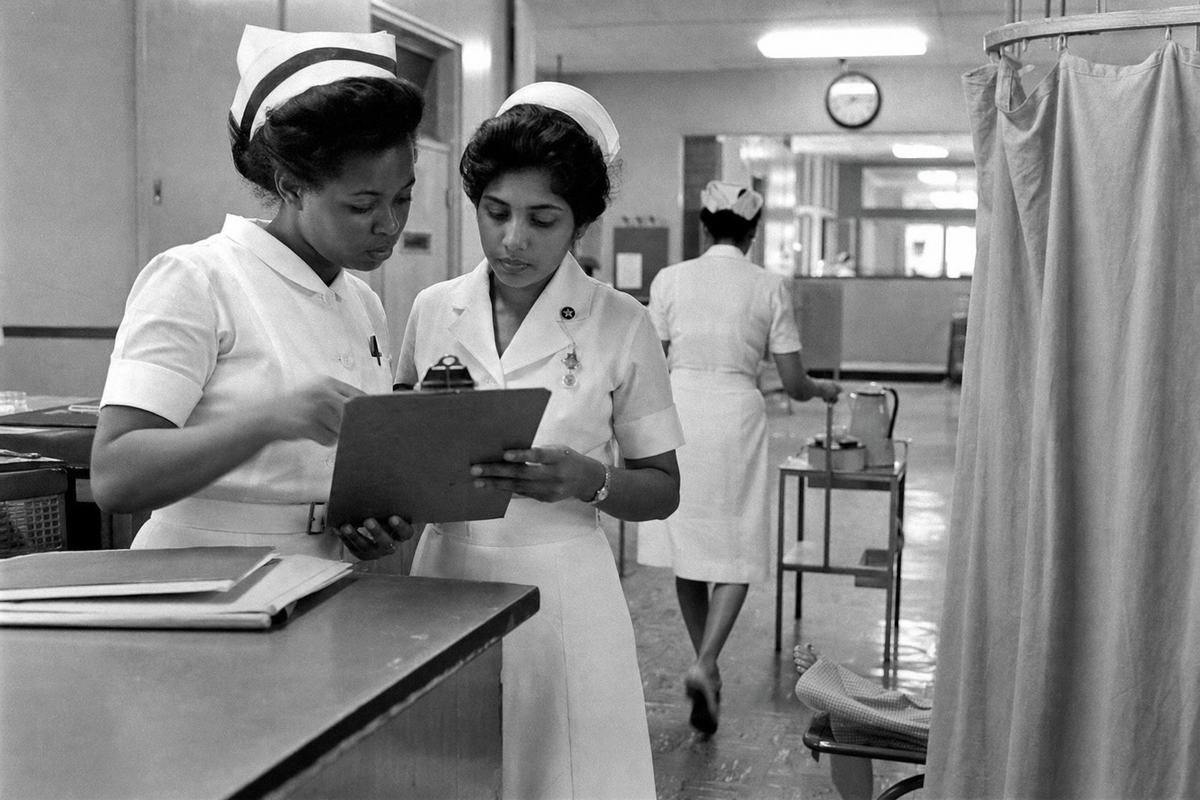 Archival black-and-white hospital corridor photograph, mid-1960s. Two nurses—one Caribbean, one South Asian—in crisp white uniforms consult a clipboard at a ward desk while a third nurse in the background wheels a trolley with enamel jugs; a patient’s slippered feet are just visible beyond a curtain. Overhead fluorescent lights create specular highlights; scuffed linoleum floor, wall clock slightly out of focus (no legible numerals). Shot on a Nikon F with 50mm lens, Kodak Tri-X 400; pronounced grain, slight motion blur in a turning hand, high-contrast highlights typical of push processing. Asymmetric composition anchored by the clipboard handover; natural skin texture and varied ages/builds.