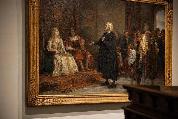 A high-resolution, museum-quality color photograph of a classical oil painting depicting Queen Isabella I of Castile receiving Christopher Columbus at the royal court, late 15th-century attire and tapestries. Shot in a quiet gallery on medium-format film (Hasselblad 500CM, Ektachrome 64) with an 80mm lens from a slight off-center angle to avoid reflections; even, gentle raking light reveals fine craquelure, subtle varnish yellowing, and brushwork around faces and hands. The gilded frame’s left edge and a strip of neutral wall are visible; a low gallery bench intrudes at bottom right, softly out of focus. No labels or signage in frame. Accurate color reproduction, natural falloff toward corners, and fine, even film grain.