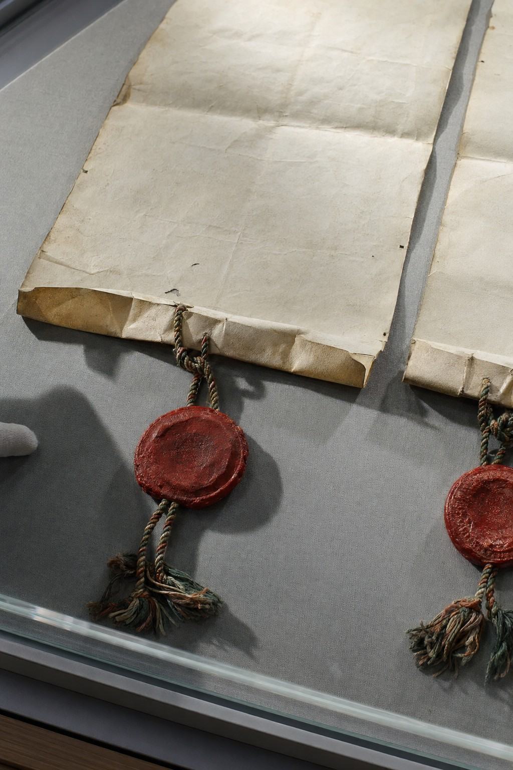 Archival object-study photograph of the 1486 preliminary capitulations: two parchment leaves under glass with wax seals suspended from braided silk cords. Shot on a copy stand with cross-polarized lights, Nikon F3 and 105mm Micro-Nikkor on Ektachrome 64; shallow raking angle from the lower left to emphasize parchment cockling, edge tears, and the relief of the red wax. Composition cropped to exclude any handwriting; only blank margins, folds, seals, and cords are shown. A grey conservation mat and the beveled acrylic case edge appear at the periphery; a faint shadow of a cotton-gloved hand falls across the mat. Clean, accurate color with minimal glare and fine film grain.