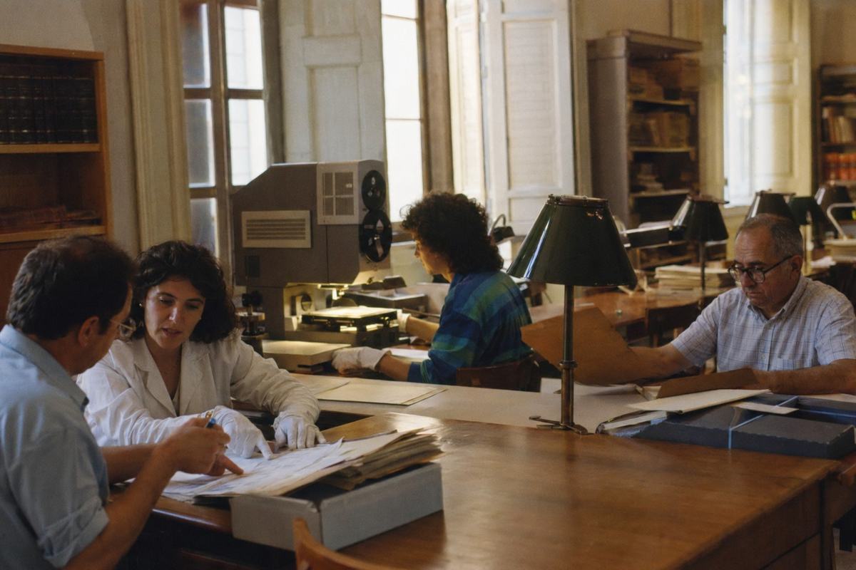 Ambient interior photograph of the Archivo General de Indias reading room in 1986: long wooden tables with green-shaded lamps, a microfilm reader and an optical-disc workstation in use. Shot on Kodachrome 64 with a 50mm lens, hand-held; mixed daylight and tungsten produce warm shadows, moderate grain, and slight edge blur. Asymmetric composition with an archivist in white cotton gloves conferring with a researcher at left; two other readers of different ages study folders at right. Tall sash windows and shelving form the backdrop. No legible text in frame; box labels turned away.