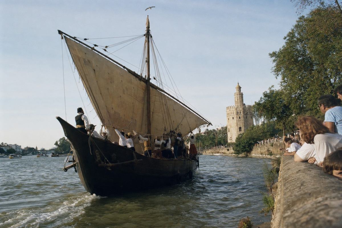 Color photograph of a caravel replica under sail on the Guadalquivir during Seville’s 1986 quincentenary regatta, crew in period garb hauling on the lateen yard. Shot on Fujicolor 100 with a Nikon FM2 and 28mm lens from the riverbank, late-afternoon sun; warm cast, fine grain, and slight motion blur in the water. Asymmetric framing with the bow cutting into the left foreground; spectators lean over the stone quay at right, Torre del Oro and plane trees receding in the background; no legible signage visible. Rigging taut, wake fanning, a gull arcs above the mast.