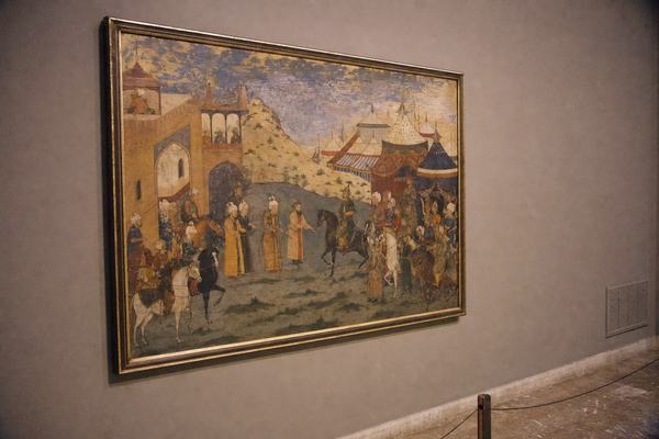 A 2001 digital press photograph taken obliquely in a museum gallery in Baghdad, showing a Timurid-style late-medieval court painting that depicts envoys meeting between Baghdad’s gates and Timur’s encampment during the 1401 parley. Camera: Nikon D1 at 50mm, f/2, ISO 800; shallow depth of field isolates the framed painting, with fine high-ISO noise and slight warm color shift. Even, naturalistic ceiling track lighting; soft falloff to a scuffed stone floor and a neutral wall. A low barrier post and a faint HVAC grille sit near the frame edge; no people present. No labels or wall text visible; the artwork itself shows no calligraphy bands or lettering. Slight barrel distortion and edge vignetting from the lens. Asymmetric composition with the painting offset to the left and empty wall space to the right.