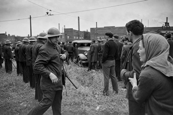 Archival black-and-white photograph, silver gelatin print from a 4x5 cellulose nitrate negative, taken with a Graflex Speed Graphic (127mm lens) at roughly f/11, 1/200s; tense police line in steel helmets facing a mixed crowd of steelworkers and neighbors outside the Republic Steel South Chicago Works gates, moments before gunfire on May 30, 1937; overcast holiday light, moderate grain, slight edge vignetting, light wind motion blur in coat hems and prairie grass; asymmetric composition with one officer stepping forward while loosening his baton strap as a woman in a headscarf in the near foreground clutches a man’s sleeve; background details include brick mill buildings, low smokestacks, a dented parked truck, weeds with an empty bottle, and a trolley wire overhead; no legible text or signage anywhere in frame.