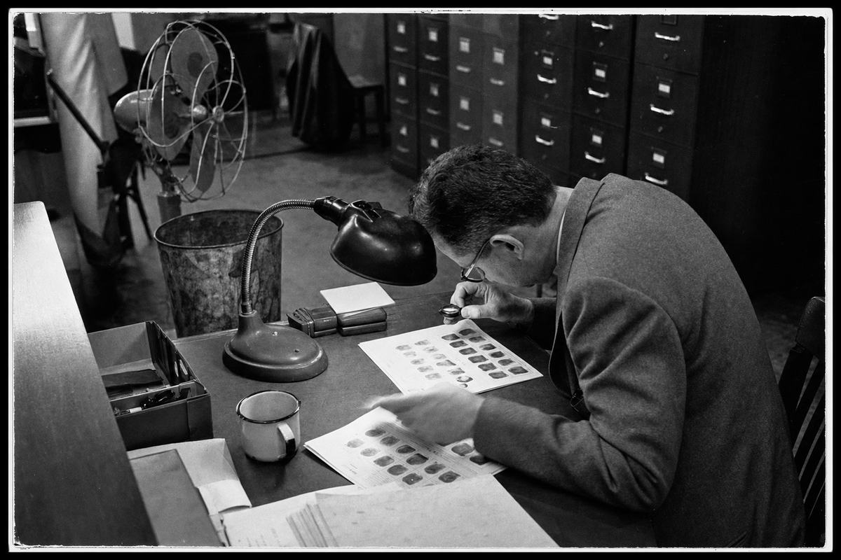 Mid-1950s black-and-white press photograph inside a Department of Justice identification division room; Kodak Tri-X 400, moderate grain and high contrast under tungsten desk lamps; shot on a Leica M3 with a 50mm lens. Asymmetric frame centered on a clerk leaning in with a loupe over two fingerprint cards under a gooseneck lamp; rows of tall steel filing cabinets recede in the background, with a pedestal fan, a dented wastebasket, and a coffee mug on a blotter. Slight motion blur in the clerk’s hand turning a card, faint edge vignetting, and contact-print border. Papers are angled away so no text or lettering is legible anywhere in frame.