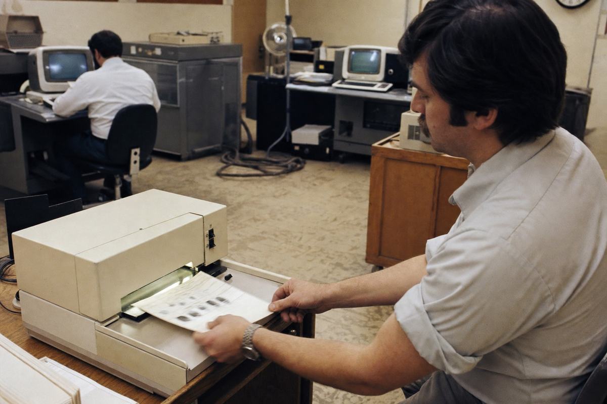 Color photograph from 1978 inside an AFIS pilot operations floor (Chicago) showing early optical fingerprint scanners and computer terminals; Kodachrome 64 with warm saturation and gentle contrast; shot on a Nikon F2 with 50mm lens. Dominant subject: a technician with rolled-up sleeves feeding a fingerprint card into a beige optical scanner; background includes two CRT terminals with soft glow (no legible screen text), coiled cables on scuffed linoleum, another operator hunched over a terminal, and a wall clock partly out of frame. Slight motion blur on the card, visible film grain, and mixed fluorescent/daylight color cast; no visible signage or lettering.