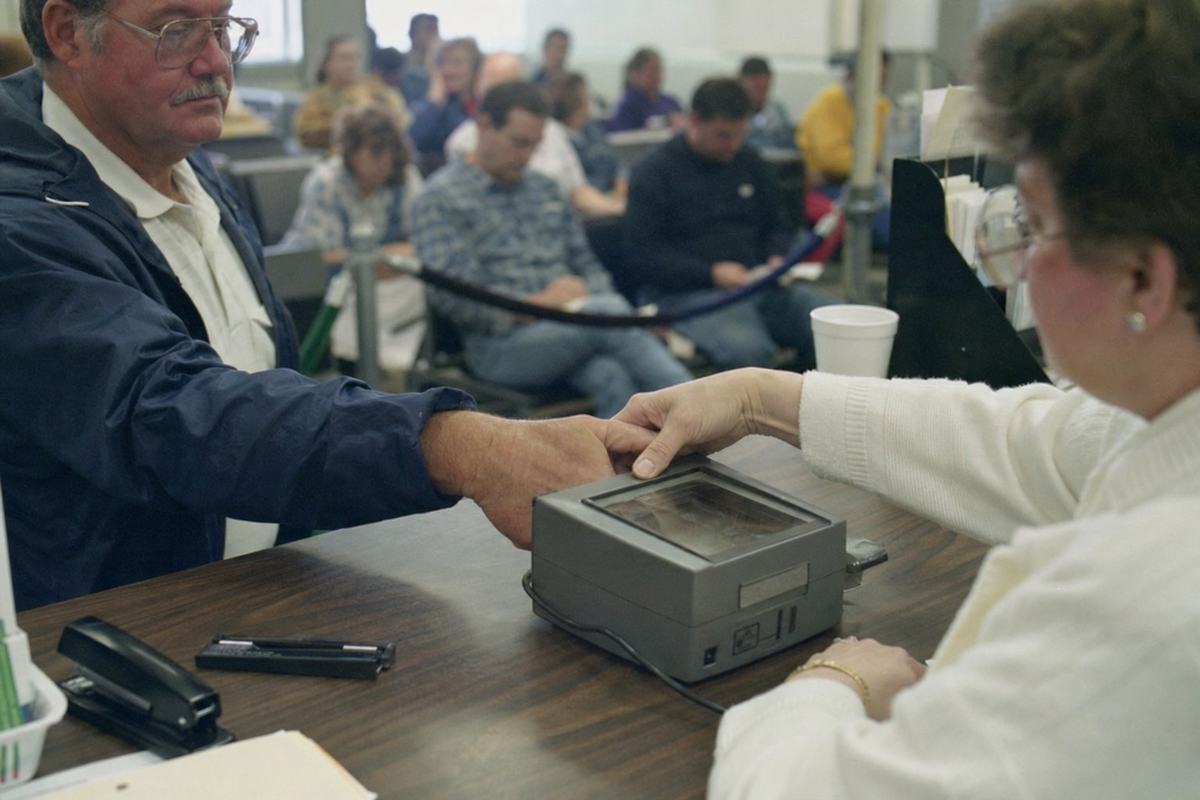 Early-1990s color photograph at a DMV enrollment counter capturing a thumbprint for a Federal Identity Card renewal; Fujicolor 400 with slight greenish cast from fluorescent lights; shot on a Canon EOS A2 with a 35mm lens, shallow depth of field. Dominant subject: a clerk’s hand guiding a middle-aged customer’s right thumb onto a glass platen of a gray desktop scanner; background shows a rope queue and seated patrons blurred, with incidental items like a stapler and a cup on the counter. Composition is candid and off-center; any posters or forms are turned away or cropped so no text or signage is legible.